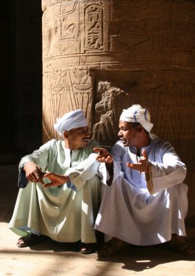 2 Maenner sitzen diskutierend am Fuß einer Saeule mit Hieroglyphen